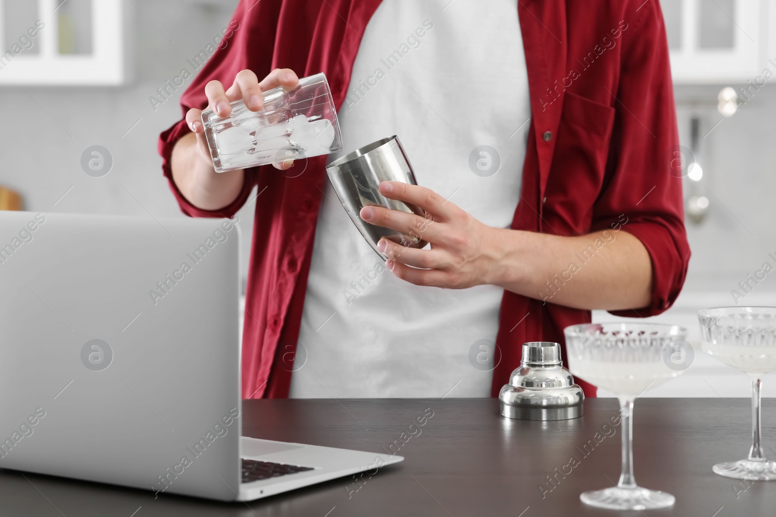 Photo of Man learning to make cocktail with online video on laptop at wooden table in kitchen, closeup. Time for hobby