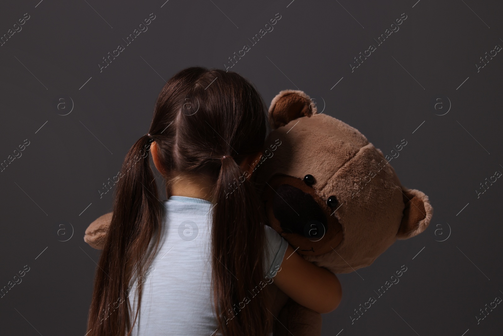 Photo of Child abuse. Upset little girl with teddy bear on gray background, back view