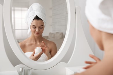 Photo of Beautiful young woman with hair wrapped in towel holding face cream near mirror at home