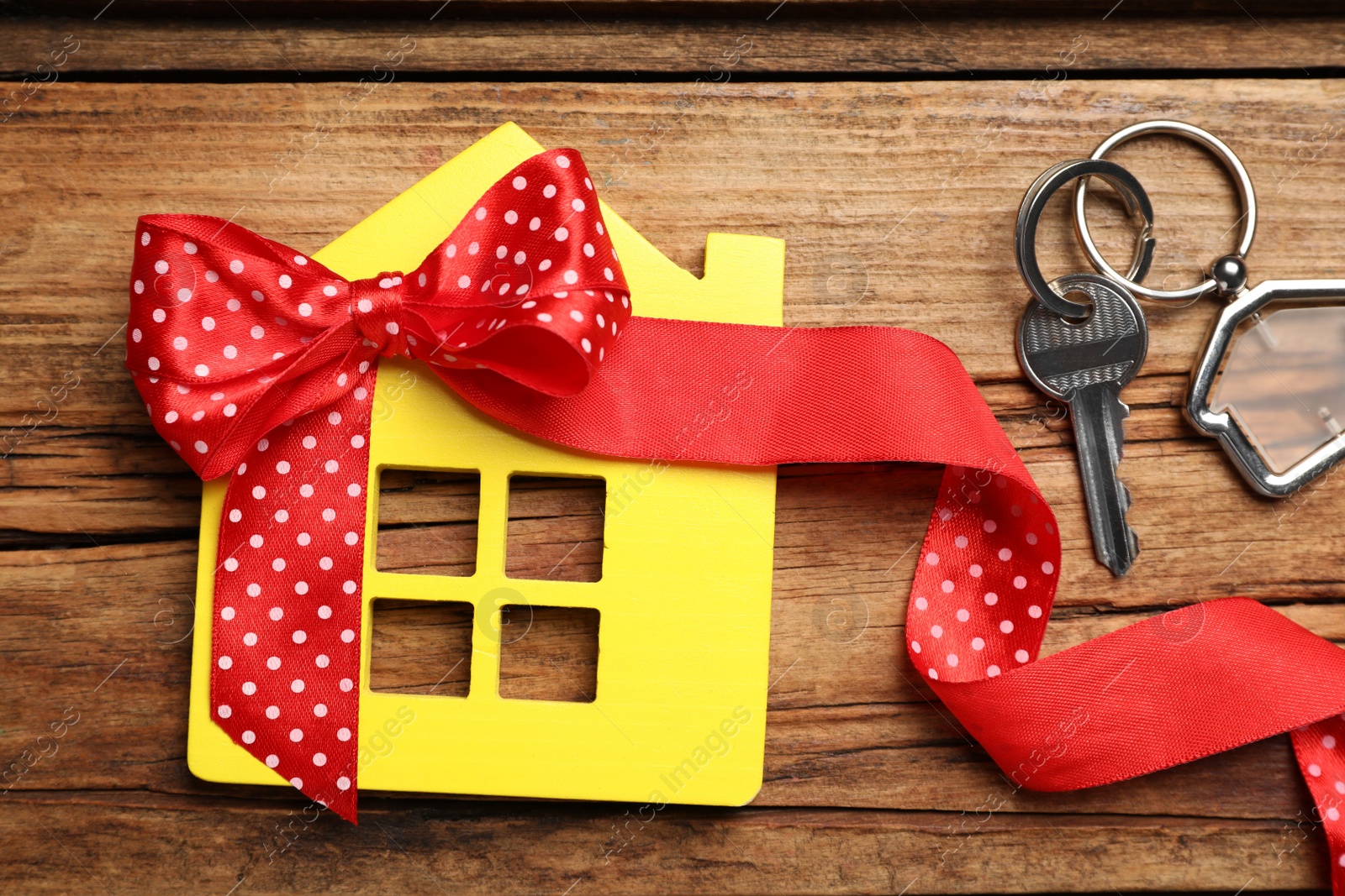 Photo of Yellow house model with bow and key on wooden table, flat lay. Housewarming party