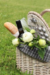 Picnic basket with wine, bread and flowers on green grass outdoors