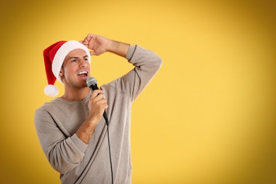 Photo of Emotional man in Santa Claus hat singing with microphone on yellow background, space for text. Christmas music