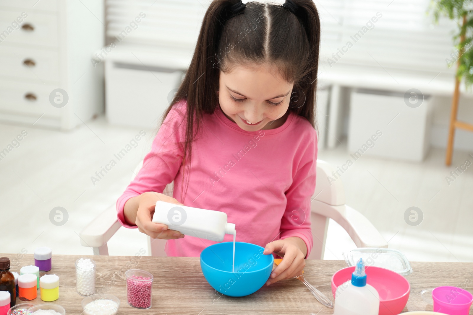 Photo of Cute little girl pouring glue into bowl at table in room. DIY slime toy