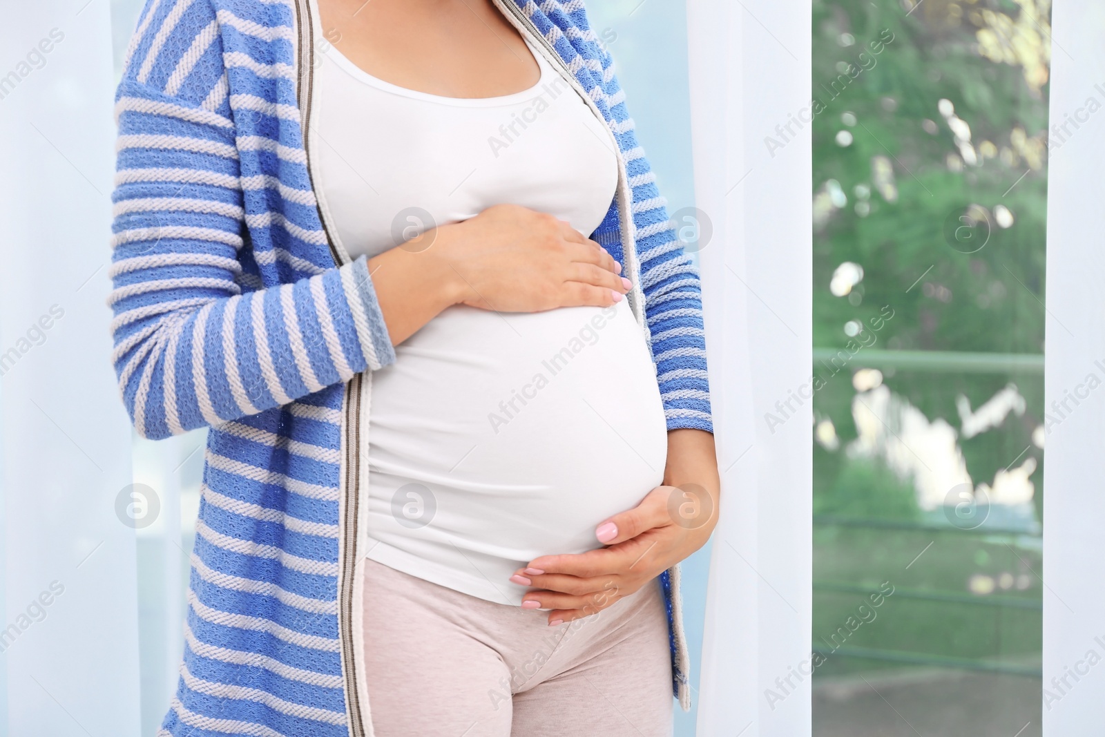 Photo of Young pregnant woman near window at home, closeup