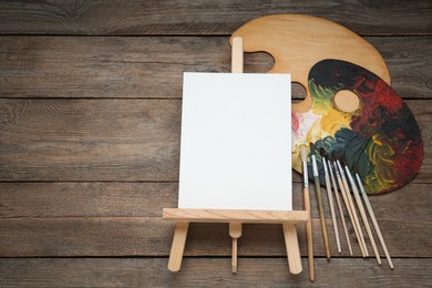 Photo of Easel with blank canvas, brushes and palettes on wooden table, flat lay. Space for text