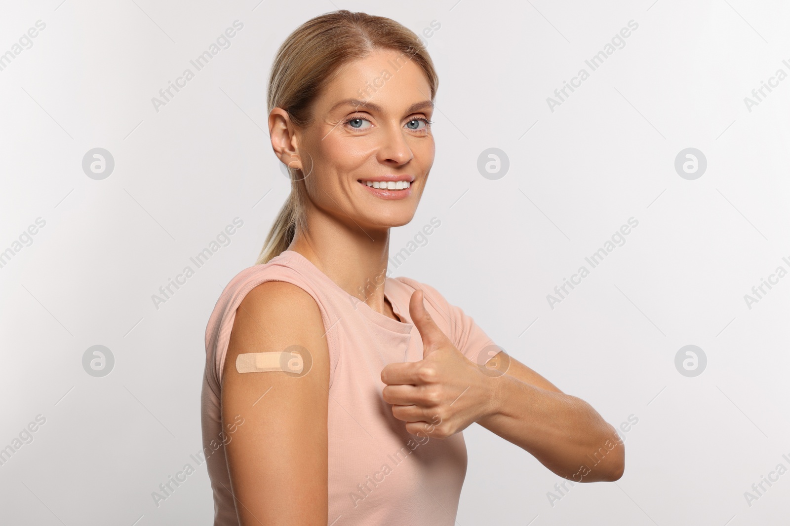 Photo of Smiling woman with adhesive bandage on arm after vaccination showing thumb up on light background