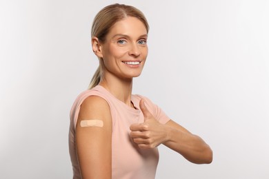 Smiling woman with adhesive bandage on arm after vaccination showing thumb up on light background