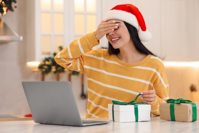 Celebrating Christmas online with exchanged by mail presents. Smiling woman covering eyes before opening gift box during video call at home
