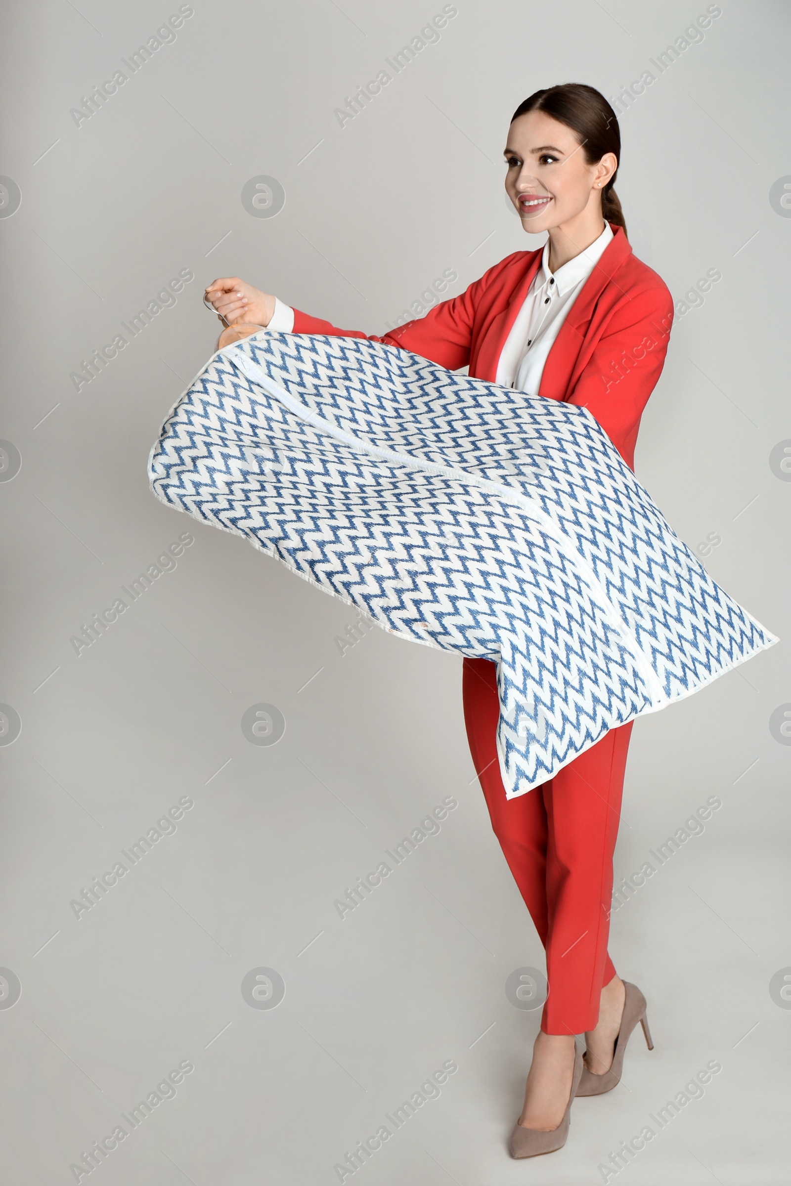 Photo of Young woman holding hanger with clothes in garment cover on light grey background. Dry-cleaning service