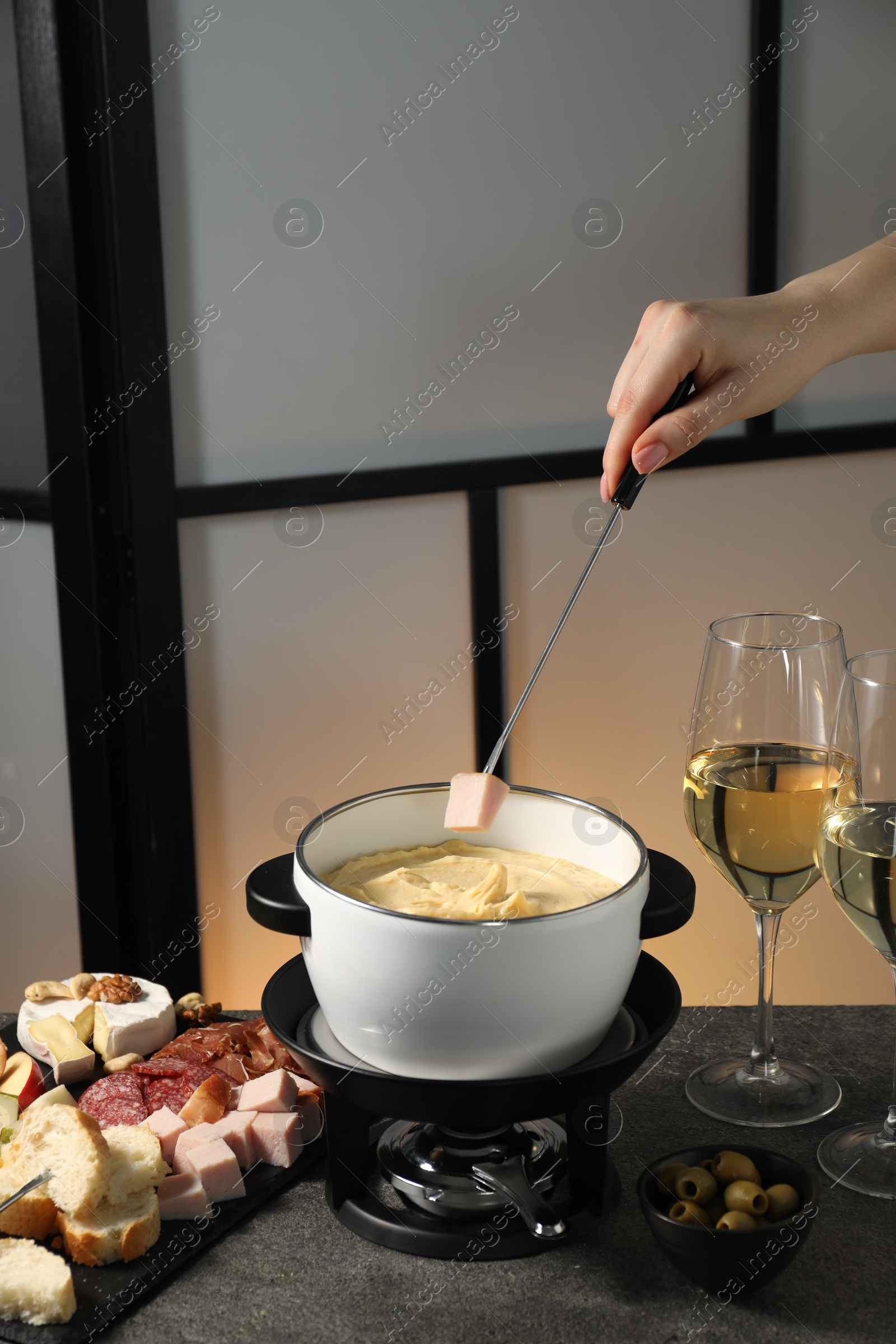 Photo of Woman dipping piece of ham into fondue pot with melted cheese at grey table with wine and snacks, closeup