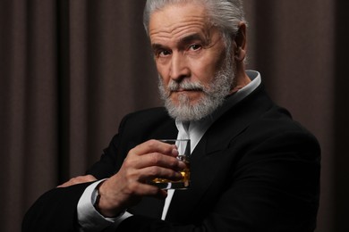 Senior man in formal suit holding glass of whiskey with ice cubes on brown background