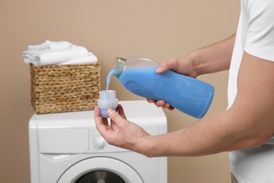 Photo of Man pouring fabric softener from bottle into cap near washing machine indoors, closeup