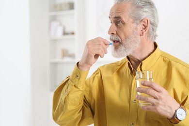 Photo of Senior man with glass of water taking pill indoors. Space for text