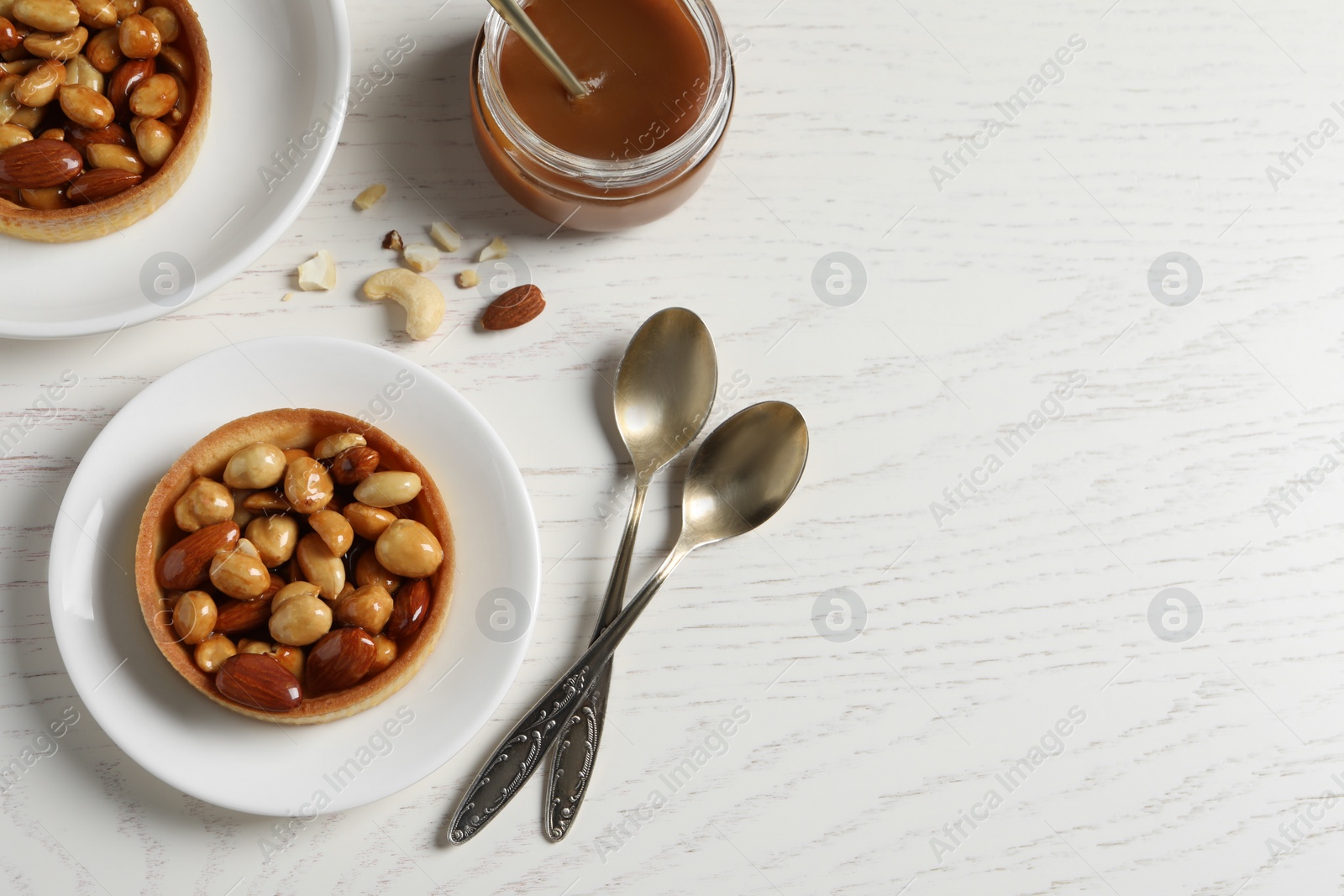 Photo of Tasty dessert. Tartlets with caramelized nuts, boiled condensed milk and spoons on white wooden table, flat lay. Space for text