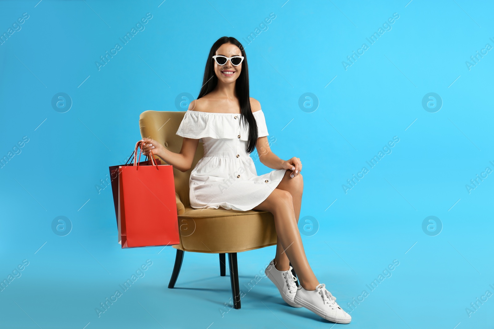 Photo of Beautiful young woman with paper shopping bags in armchair on light blue background