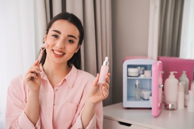 Woman doing face massage at dressing table with cosmetic refrigerator indoors