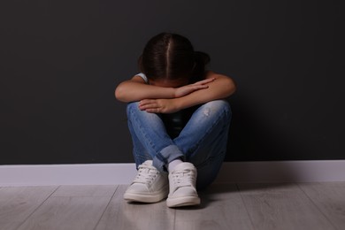 Photo of Child abuse. Upset little girl sitting on floor near gray wall indoors