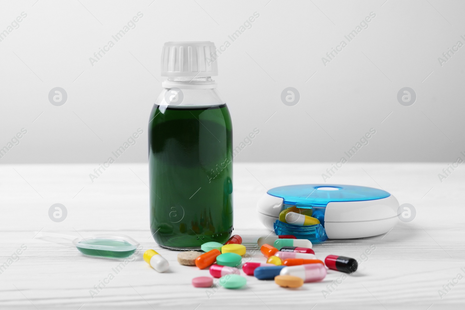 Photo of Bottle of syrup, dosing spoon, weekly pill organizer and pills on white table against light grey background. Cold medicine