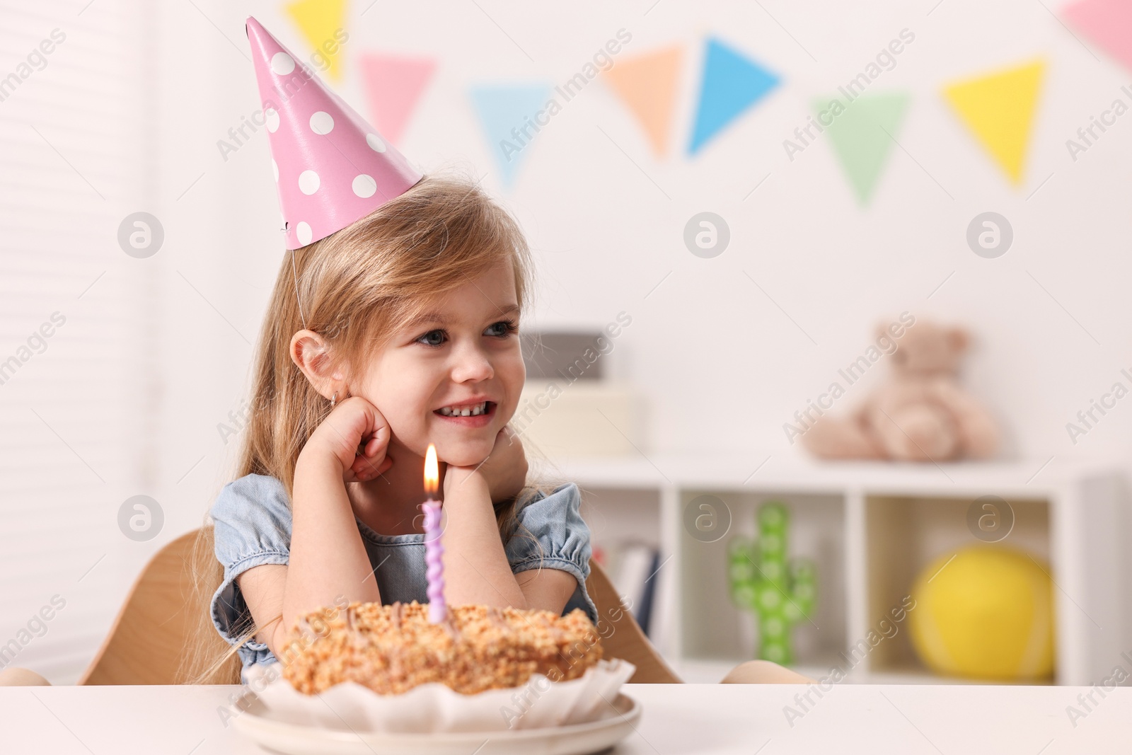 Photo of Cute girl in party hat with birthday cake at table indoors, space for text