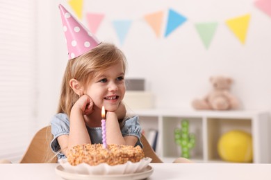 Photo of Cute girl in party hat with birthday cake at table indoors, space for text