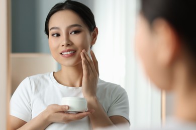 Happy woman applying face cream near mirror at home