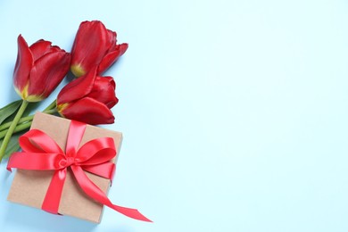 Photo of Beautiful gift box with bow and red tulips on light blue background, flat lay. Space for text