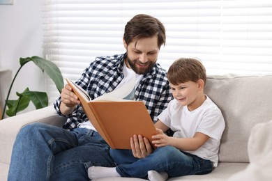 Photo of Happy dad and son reading book together on sofa at home