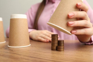Photo of Shell game. Woman showing stack of coins under cup at wooden table, closeup