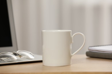 Photo of White ceramic mug, glasses and laptop on wooden table at workplace