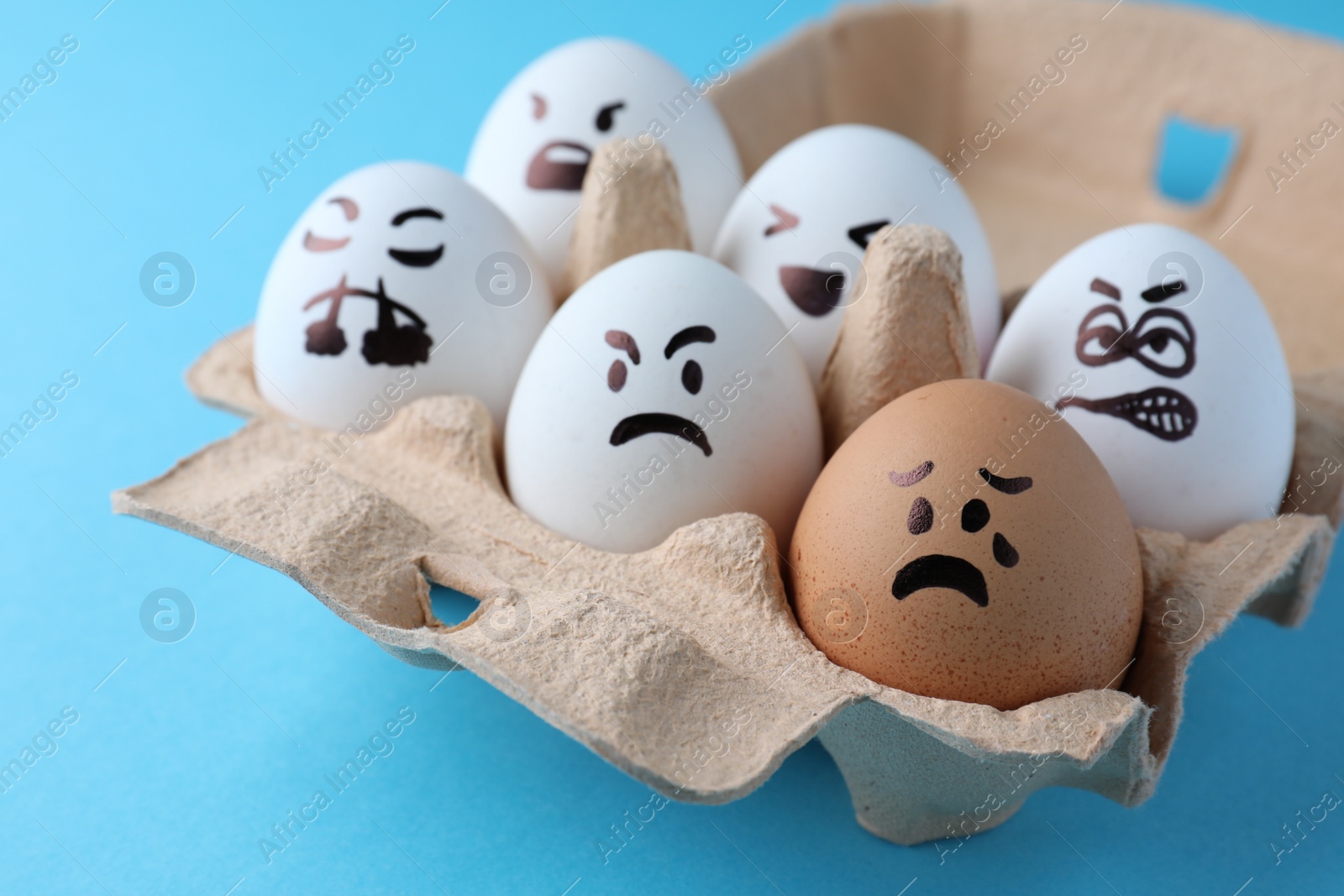 Photo of Brown egg with upset face among aggressively disposed white ones in carton box on turquoise background, closeup. Bullying concept