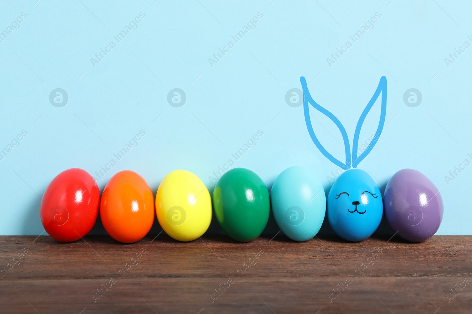 Image of One egg with drawn face and ears as Easter bunny among others on wooden table against light blue background