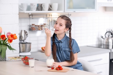 Cute girl eating tasty yogurt at table in kitchen