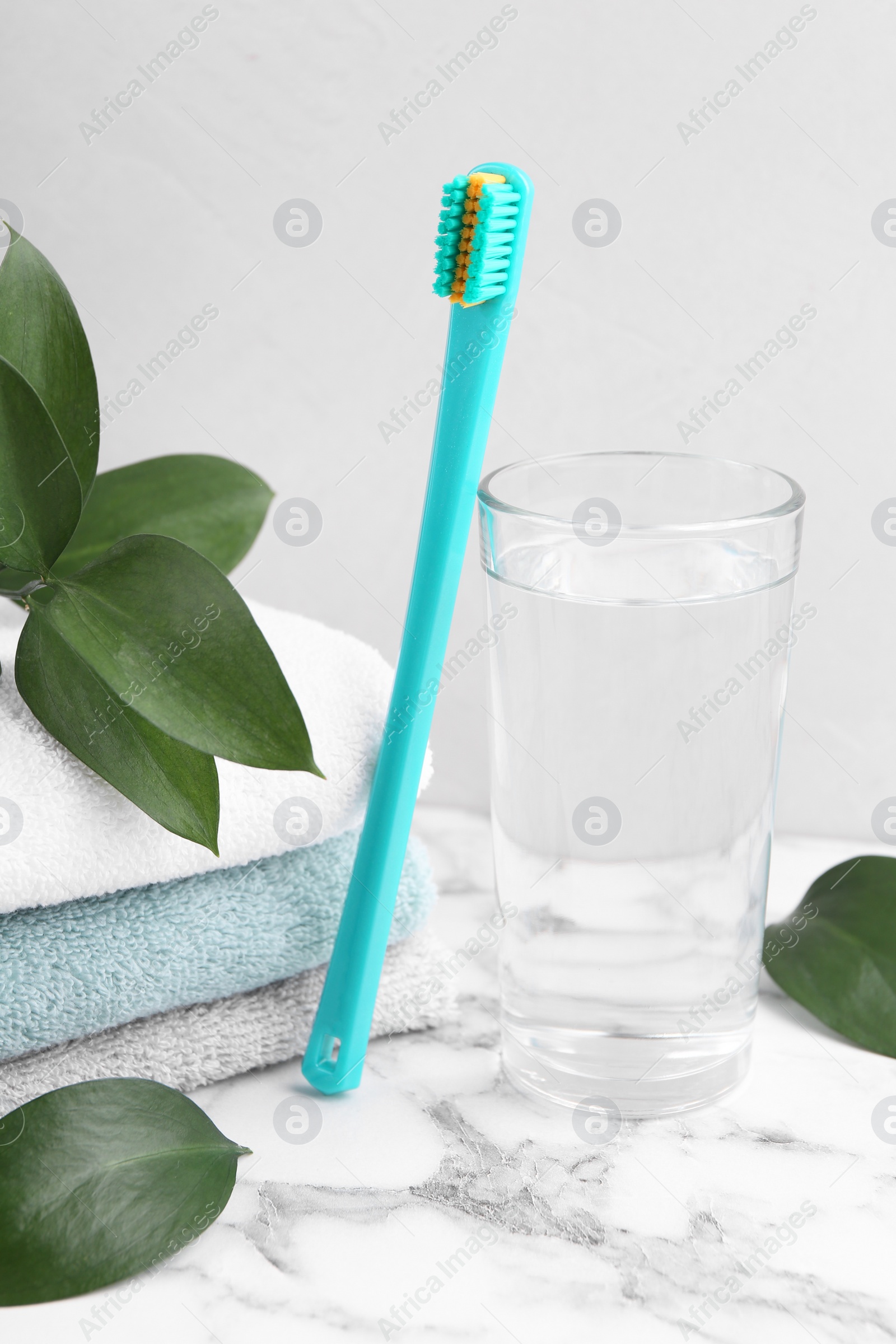 Photo of Plastic toothbrush, glass of water, towels and green leaves on white marble table