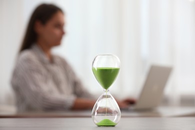 Photo of Hourglass with light green flowing sand on table. Woman using laptop indoors, selective focus
