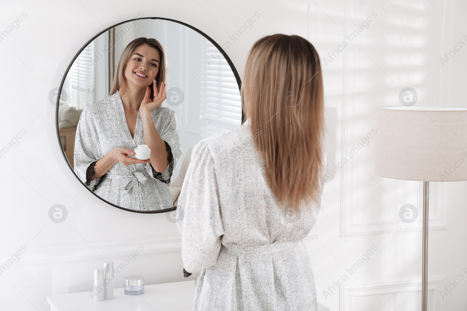 Photo of Beautiful young woman applying cream near mirror in room