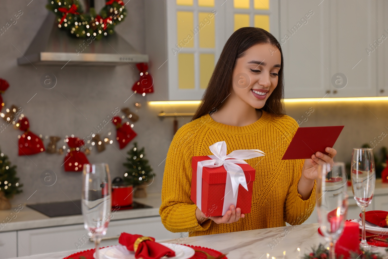 Photo of Happy young woman with Christmas gift reading greeting card at table in kitchen