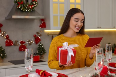 Photo of Happy young woman with Christmas gift reading greeting card at table in kitchen