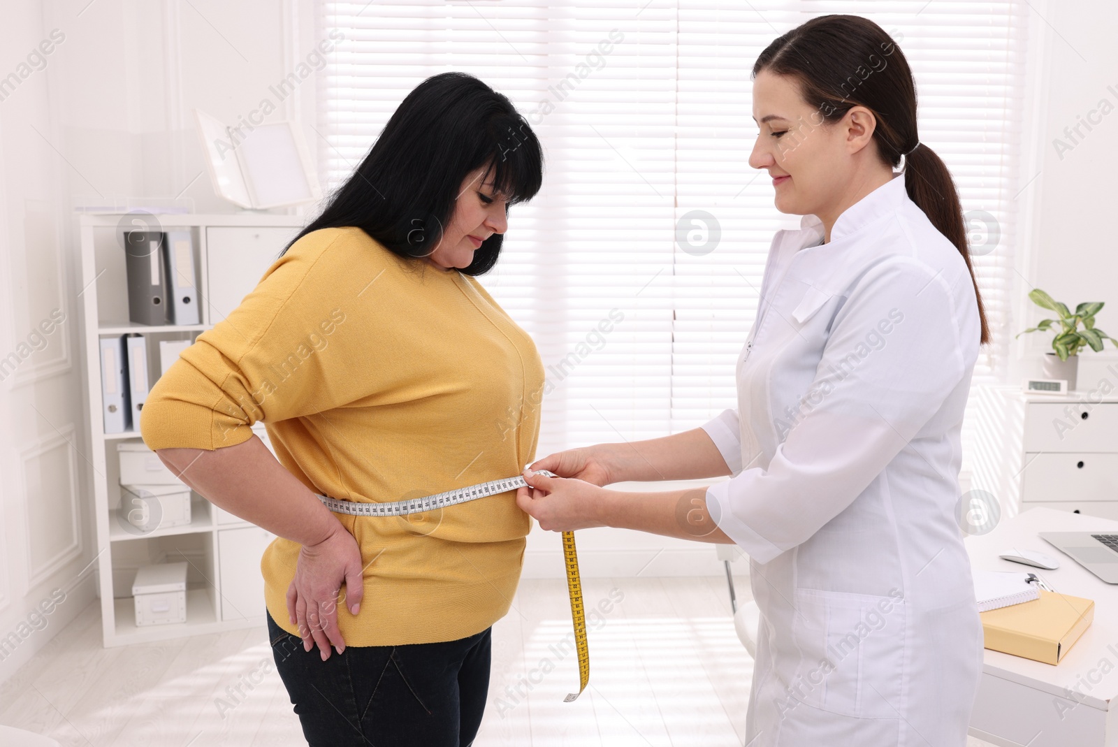 Photo of Nutritionist measuring overweight woman's waist with tape in clinic
