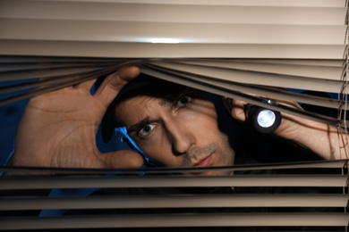 Male security guard with flashlight looking through window blinds in darkness