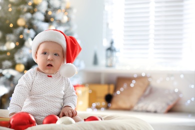 Photo of Little baby in Santa hat playing with Christmas balls indoors