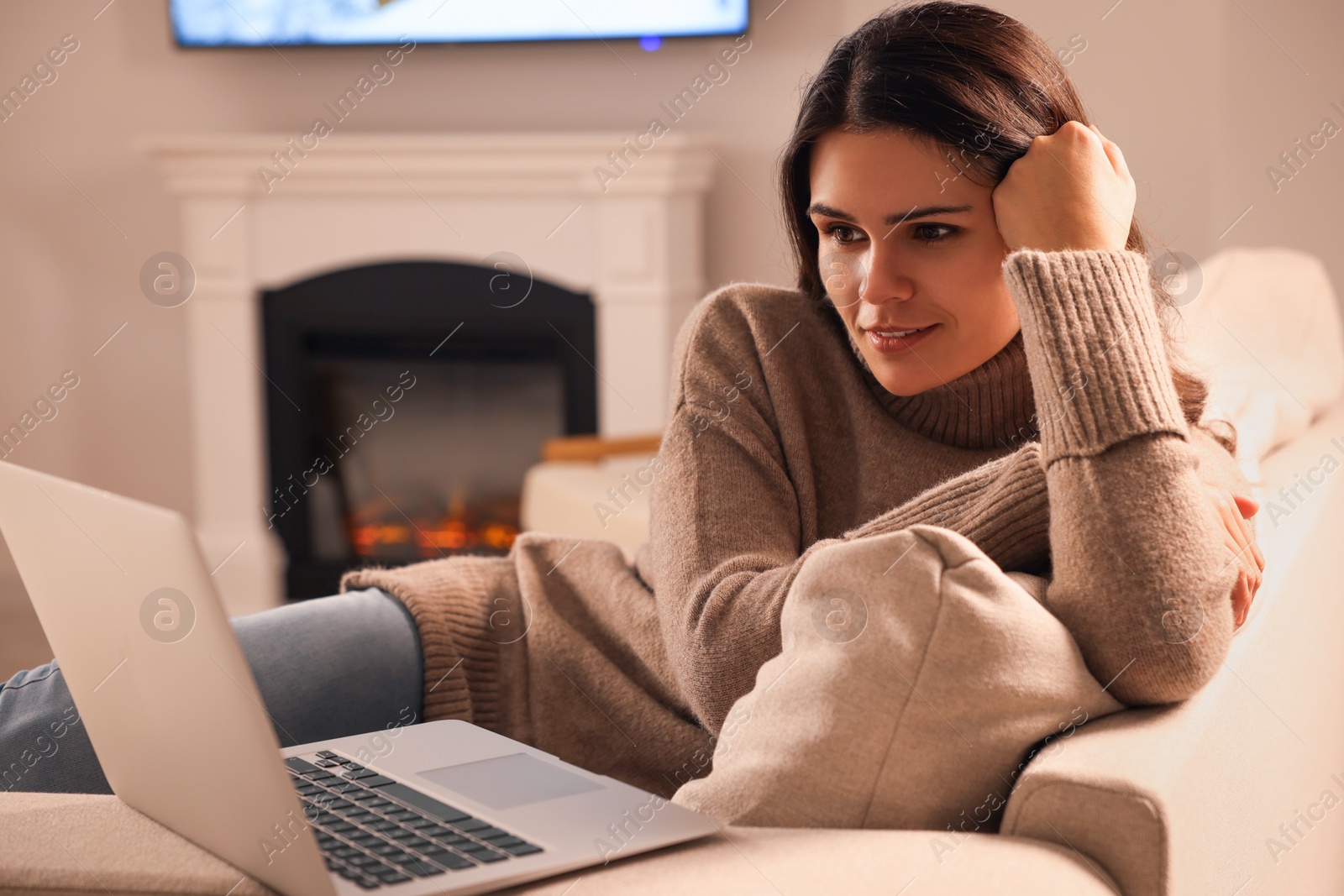 Photo of Young woman with laptop on sofa near fireplace at home