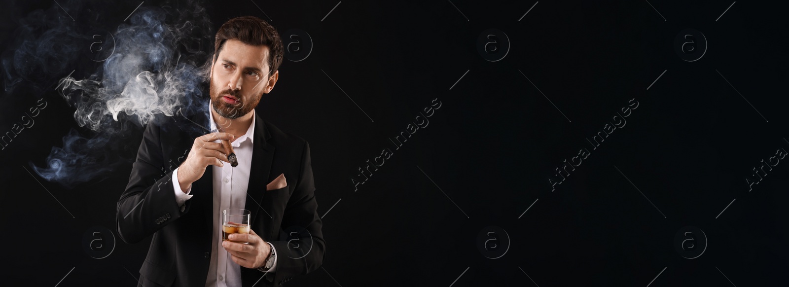 Photo of Handsome man in elegant suit with glass of whiskey smoking cigar on black background