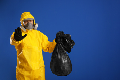 Photo of Man in chemical protective suit holding trash bag on blue background, space for text. Virus research