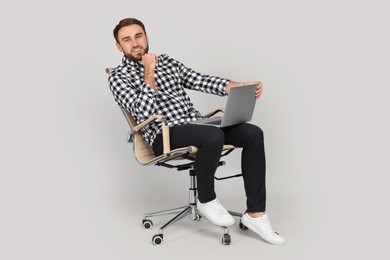Young man with laptop sitting in comfortable office chair on grey background