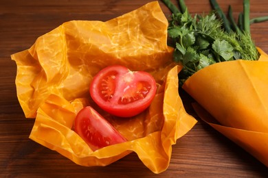 Photo of Slices of fresh tomato and herbs in beeswax food wraps on wooden table, closeup