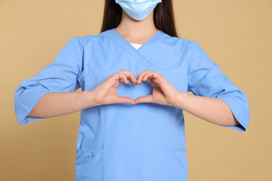 Photo of Nurse wearing medical uniform making heart with hands on light brown background, closeup