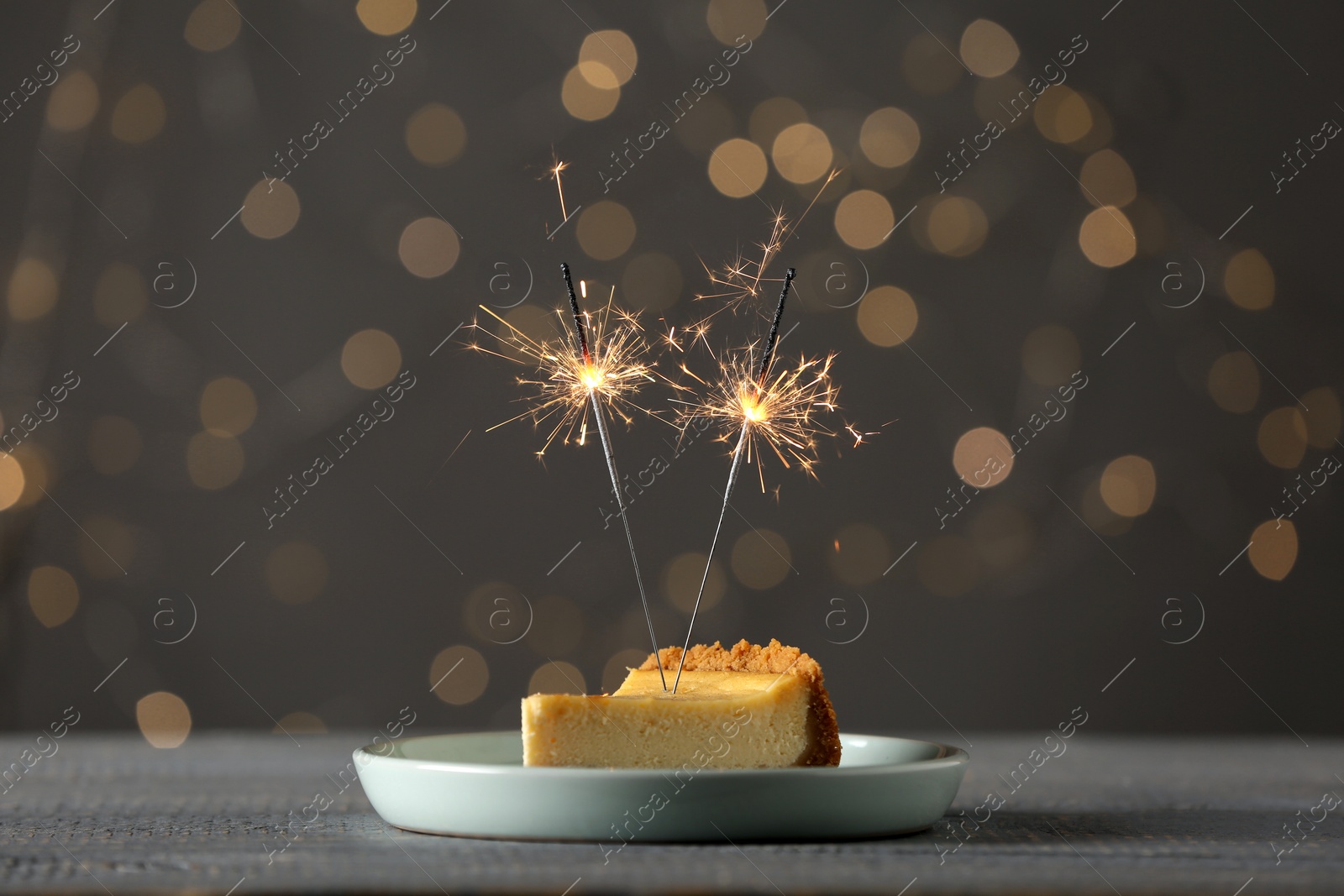 Photo of Piece of delicious cake with burning sparklers on grey wooden table against blurred lights