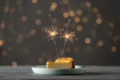 Photo of Piece of delicious cake with burning sparklers on grey wooden table against blurred lights