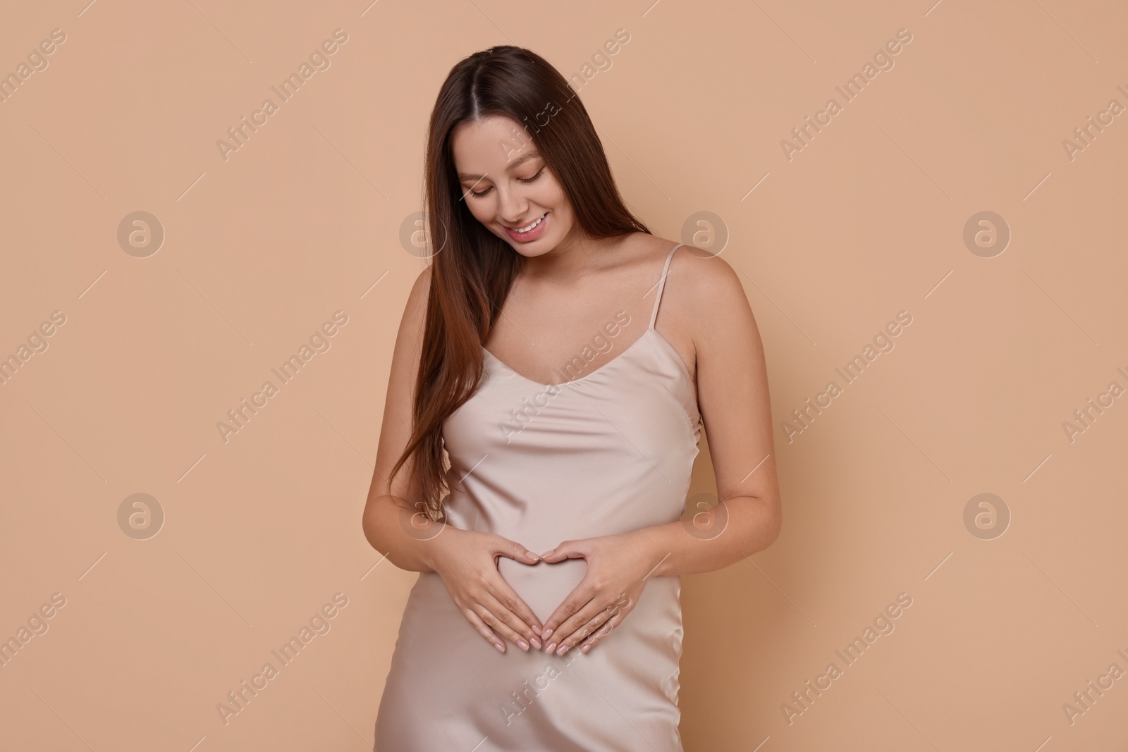 Photo of Beautiful pregnant woman in dress making heart with hands on beige background