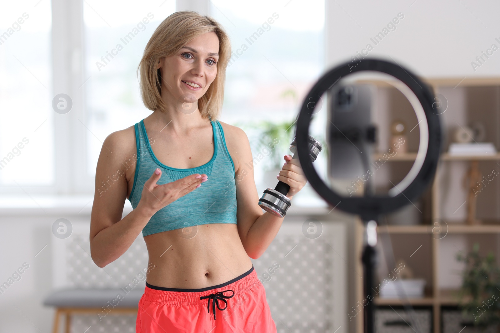 Photo of Smiling sports blogger holding dumbbell while streaming online fitness lesson with smartphone at home